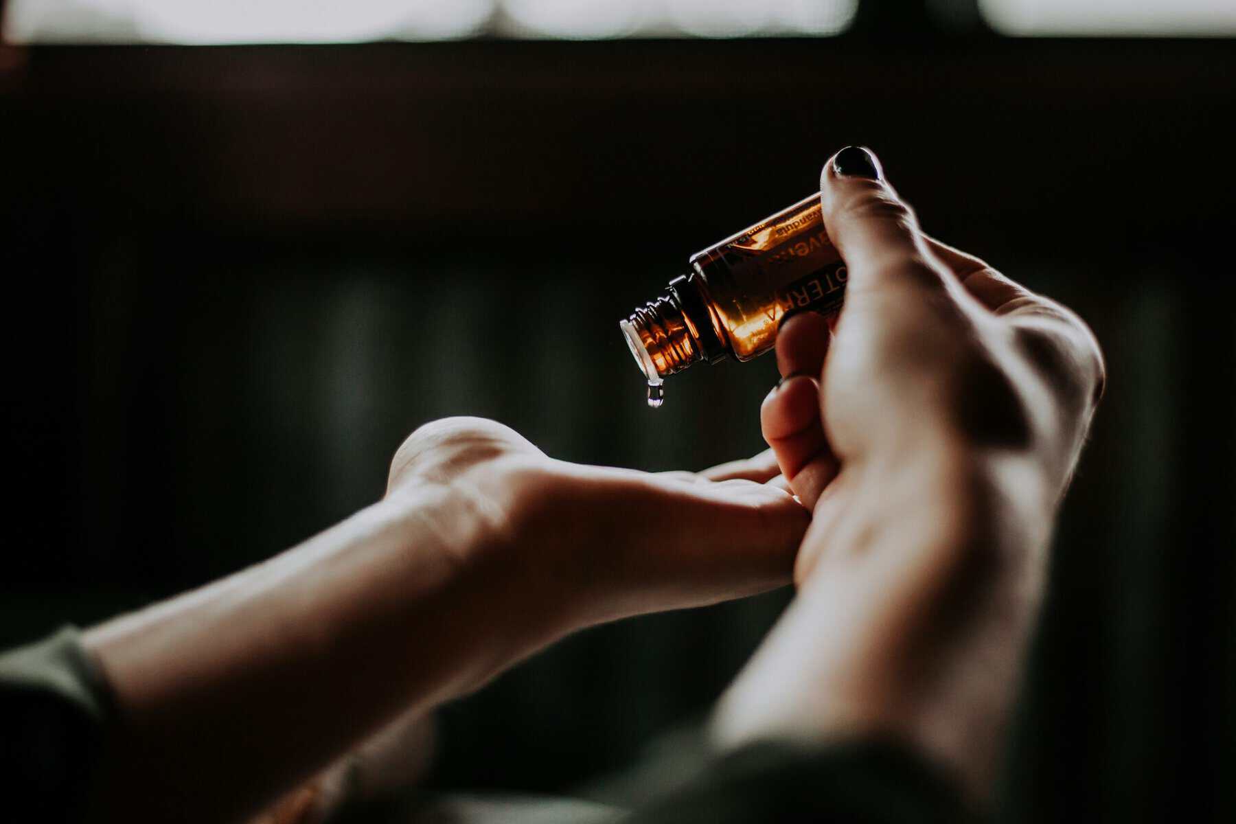 Close-up of a hand holding a small amber glass bottle, releasing a single droplet from a dropper against a dark blurred background.