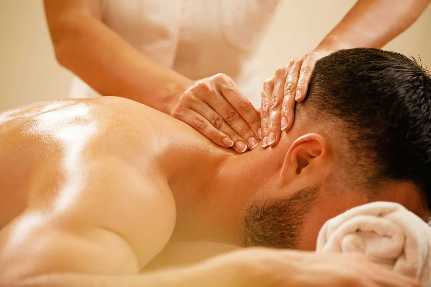 Close-up of hands massaging a person's neck and upper back as they lie on a massage table, warm lighting nearby.
