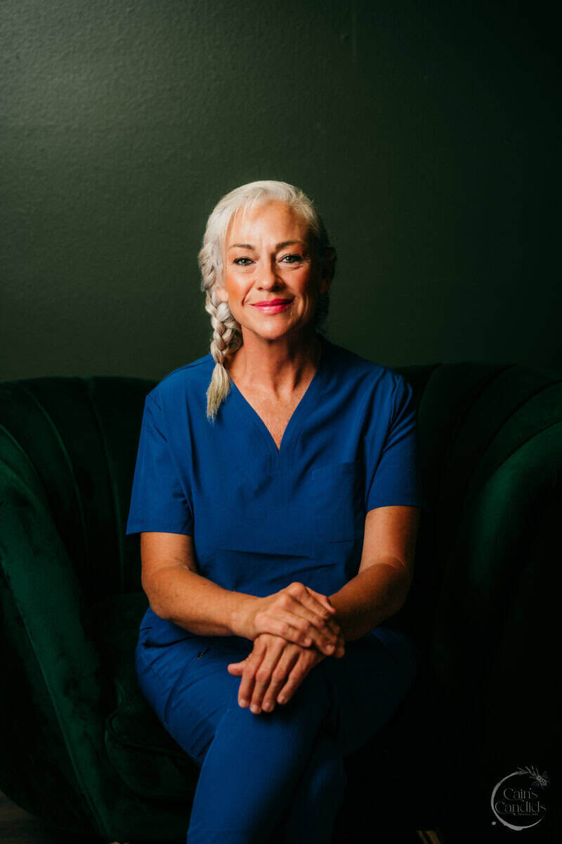 Smiling older woman with white braided hair in blue scrubs seated on a dark green sofa against a green wall.